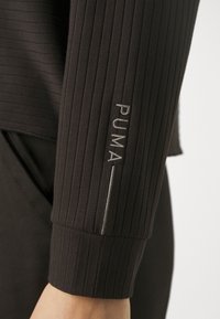 Schwarzes Langarmoberteil mit vertikal gerippter Textur. Verfügt über einen gefütterten Ärmel mit dem in Silber gestickten "PUMA"-Logo an der Seite.