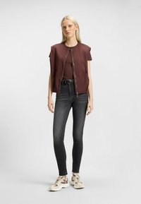 Maroon padded vest with shoulder caps, zip front, and side pockets, worn over a dark top, paired with black skinny jeans and beige sneakers.