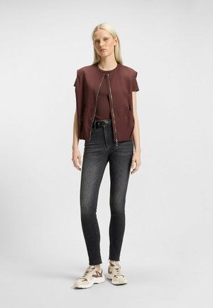 Gilet imbottito color borgogna con spalline, chiusura con zip e tasche laterali, indossato sopra un top scuro, abbinato a jeans skinny neri e sneakers beige.