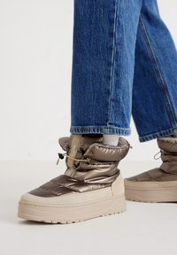 Pūšļaini, līdz teļam augsti zābaki ar spīdīgu bronzas materiālu un krēmkrāsas zoli. Iekļauj saites aizdari un teksturētu augšdaļu. Saskaņoti ar ziliem džinsiem.