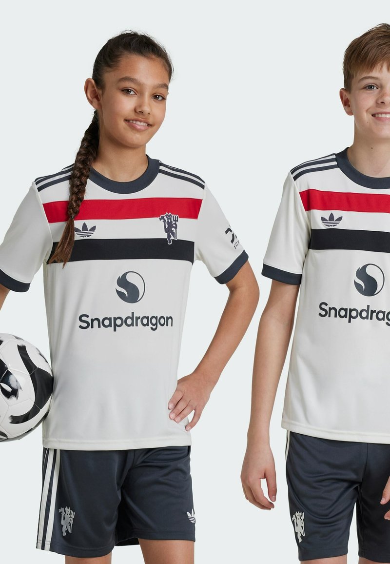 Maillots de sport blancs avec des rayures rouges et noires, associés à des shorts foncés. Comprend des logos et une finition en tissu texturé.