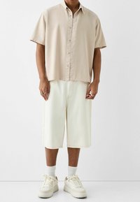 Beżowa koszula na krótki rękaw z guzikami, jasne spodenki do kolan oraz kremowe trampki z białymi skarpetkami. Gładka tekstura i luźne dopasowanie.