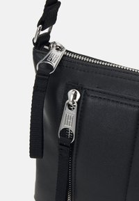 Zwarte leren crossbody tas met zilveren hardware, voorzien van dubbele ritsen en een gestructureerd frontpaneel. Inclusief een verstelbare webbing schouderband.