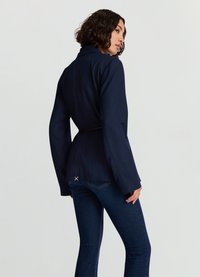 Veste bleu marine à coupe ajustée, avec une ceinture à la taille, des manches longues et une fente au dos. Associée à un jean denim foncé.