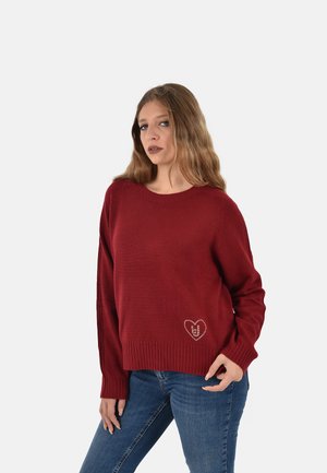 Maglione rosso lavorato a maglia con maniche lunghe, orlo a coste e un design a forma di cuore sul davanti. Abbinato a jeans blu. Texture morbida.