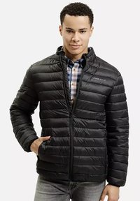 Czarna pikowana kurtka z poziomym wzorem, z zamkiem z przodu, stójką i subtelnym logo. Noszona na wierzchu koszuli w kratę.