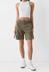 Khaki cargo šortky s několika bočními kapsami, volným střihem a knoflíkovým zapínáním, v kombinaci s bílým žebrovaným tílkem a bílými teniskami.