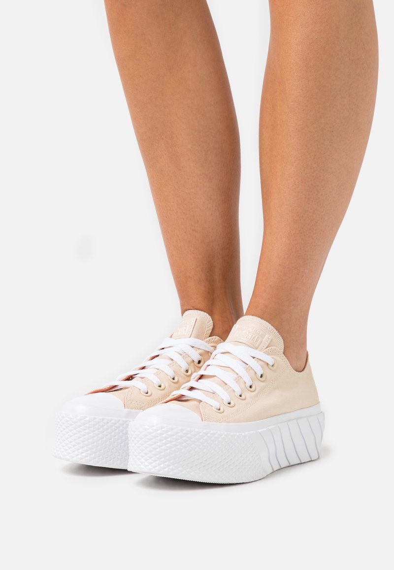Jambes portant des baskets à plateforme beige avec des lacets blancs et des semelles épaisses blanches texturées, debout sur un fond clair uni.