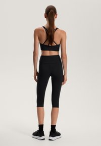 Czarny sportowy stanik z krzyżującymi się ramiączkami, w zestawieniu z czarnymi legginsami do kolan i czarnymi tenisówkami. Gładki materiał, dopasowany krój.