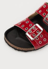 Sandale en vernis rouge avec deux larges brides et boucles en argent, dotée de découpes circulaires, montée sur une semelle noire texturée.