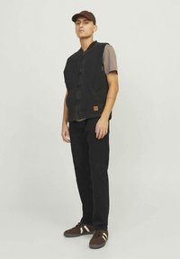 Chaleco de mezclilla negro con cremallera en el frente y bolsillos laterales, usado sobre una camisa marrón. Combinado con pantalones negros y zapatillas marrones con rayas blancas.
