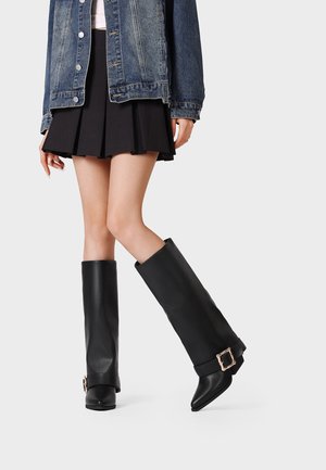 Persona che indossa una gonna nera plissettata, una giacca di jeans e stivali neri al ginocchio con grandi fibbie dorate su uno sfondo chiaro e semplice.