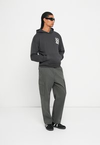 Άνδρας με πλεγμένα μαλλιά που φοράει μαύρα γυαλιά ηλίου, σκούρο γκρι φούτερ με κείμενο "Hollister 22", φαρδιά cargo παντελόνια και μαύρα αθλητικά παπούτσια.