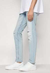 Ljusblå, slim-fit jeans i denim med slitna knädetaljer och subtila urtvättningseffekter, tillsammans med vita sneakers.