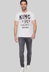 Weißes Baumwoll-T-Shirt mit dem Grafikdesign "KING of SPICY", kombiniert mit grauen Jeans und weißen Turnschuhen. Das Model steht lächelnd vor einem neutralen Hintergrund.