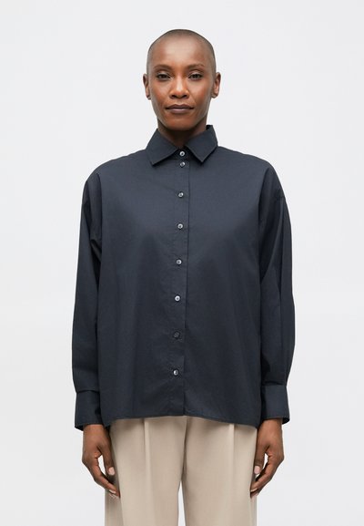 Personne portant une chemise à boutons ample de couleur foncée et un pantalon beige clair, se tenant devant un fond blanc uni.