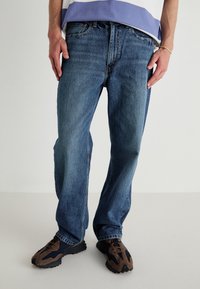 Modré džíny z denimu se střihem rovného střihu, s mírným vyblednutím a klasickým pětiprvkovým stylem kapes, doplněné o tmavě hnědé a námořnicky modré tenisky.