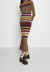 Abito a maniche lunghe a coste con strisce verticali multicolori in bordeaux, rosa, verde e giallo. Orlo svasato abbinato a scarpe con tacco nero.