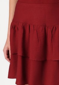 Partie d'une jupe rouge à volants avec un détail de tissu froncé, montrée sur la partie inférieure du torse et la main d'une personne.