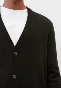 Man i en svart knappad cardigan över en vit t-shirt med rund halsringning mot en enkel ljus bakgrund.