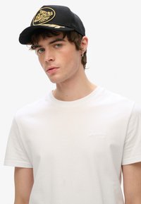 Casquette de baseball noire avec un logo doré, associée à un t-shirt blanc basique à col rond. La casquette a une visière plate et des détails brodés.