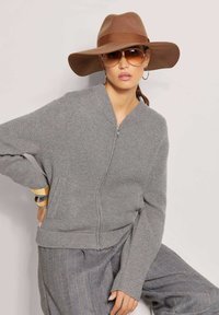Grauer Reißverschlusscardigan mit geripptem Muster und seitlichen Taschen, kombiniert mit einem braunen Hut mit breiter Krempe und großen Sonnenbrillen, vor einem hellen Hintergrund.