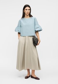 Femme portant un chemisier à manches bouffantes bleu clair, une jupe midi beige, des ballerines marron, tenant un sac à main noir matelassé.