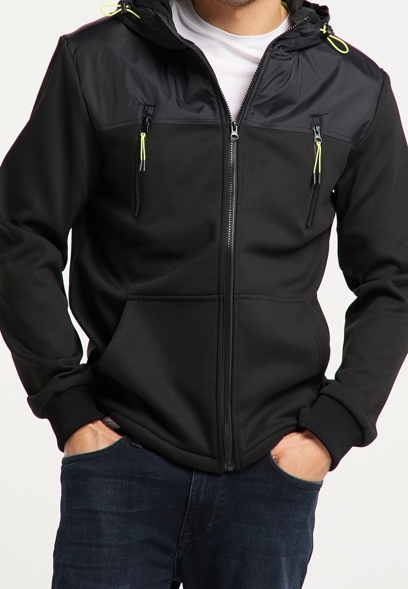 Mężczyzna w czarnej kurtce zapinanej na zamek z kapturem, z neonowożółtymi uchwytami zamka, trzymający ręce w kieszeniach, w ciemnoniebieskich dżinsach.