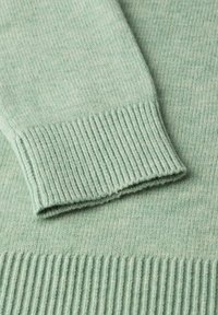 Svetle zelená pletená mikina s žebrovanými manžetami a lemem, která ukazuje jemnou texturu a subtilní barevné variace v celé látce.
