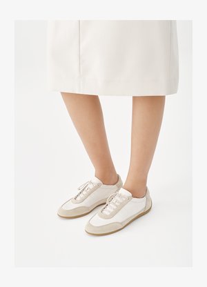 Henkilö, jolla on valkoiset tennarit, joissa on beigeä mokkanahkaa, ja valkoinen polven mittainen hame, seisomassa valkoista taustaa vasten.