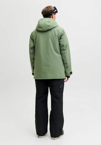 Grüne wasserdichte Jacke mit Kapuze, kombiniert mit schwarzen Hosen mit Reißverschlüssen an den Knöcheln; glatte Textur und praktische Designelemente.