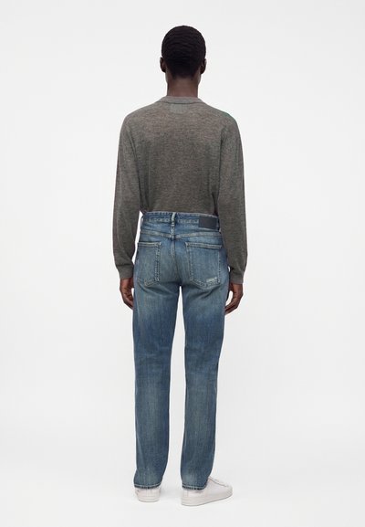 Personne portant une chemise grise à manches longues, un jean bleu et des baskets blanches, debout de dos devant un fond clair uni.