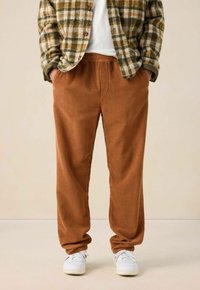Personne portant un pantalon en velours côtelé marron, des baskets blanches, une chemise blanche et une veste à carreaux verts et beiges avec les mains dans les poches.