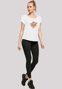 Weißes kurzärmeliges T-Shirt mit einem bunten Grafikdruck auf der Vorderseite, kombiniert mit schwarzen Leggings und schwarzen Sportschuhen.