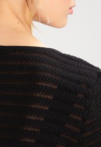 Black knitted garment featuring a textured pattern with alternating open and solid stripes. The neckline is a simple crew design.