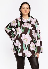 Bloemige blouse in diepbruin met grote roze pioenroosafdrukken, zachte stof, kettinghalslijn en een relaxte pasvorm, gecombineerd met zwarte slim-fit broek.