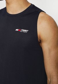 Czarna bezrękawnikowa koszulka bawełniana z małym białym i czerwonym logo "TOMMY HILFIGER" na piersi, z okrągłym dekoltem i gładką teksturą.