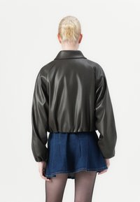 Mørkegrønn, skinnende faux skinnjakke med løs passform og vide ermer, kombinert med et blått denim miniskjørt. Vist bakfra.