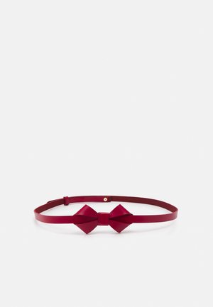 Wąski czerwony skórzany pasek z centralnym detalem w kształcie kokardy i małą okrągłą złotą sprzączką, ułożony płasko na białym tle.