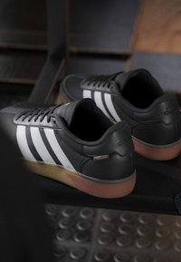 Zwarte sneakers met witte strepen en rubberen zolen die rusten op een donkere ondergrond, gedeeltelijk gereflecteerd in een nabijgelegen spiegel.