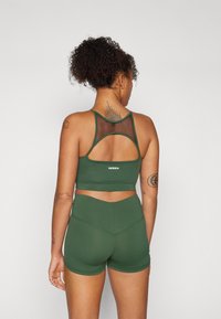 Temno zelena športna modrca z mrežastim detajlom in izrezanim hrbtom, v kombinaciji s kratkimi hlačami. Mehka, elastična tkanina, prilegnjen stil, minimalno blagovno znamčenje.