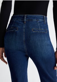 Jeans in denim blu scuro con vita alta, dotati di due tasche posteriori e cuciture a contrasto. Texture liscia con una forma aderente.