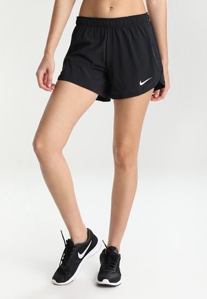 Kvinde iført sorte Nike træningsshorts og sorte Nike løbesko, stående på en hvid baggrund.