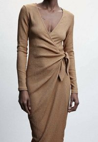 Femme portant une robe portefeuille à manches longues, beige, avec un décolleté en V profond et un nouage sur le côté, montrant un tissu texturé et une silhouette ajustée.