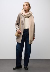 Beige uld-blend frakke over en cremefarvet sweater; parret med mørkeblå bukser med brede ben og sorte ankelstøvler. Lys beige tørklæde med frynser.