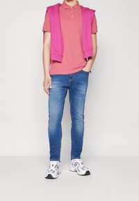 Rosa polo skjorta kombinerad med en rosa tröja draperad över axlarna, blå denimjeans och vita sportskor med svarta accenter.