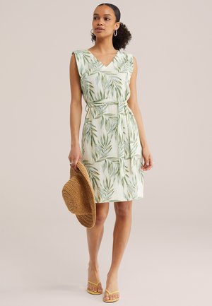 Naine kannab varrukateta põlvpikkust kleiti rohelise lehemustriga, hoiab käes kudumiga päikesevarju ja kannab kollaseid sandaale.