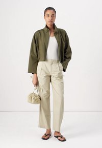 Chaqueta corta verde oliva, camiseta blanca sin mangas, pantalones beiges y chanclas negras. Llevando una pequeña bolsa de cuerda de color crema.