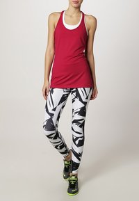 Mujer vestida con una camiseta sin mangas roja, leggings de patrón en blanco y negro, y zapatillas deportivas negras con verde neón, de pie con una pierna cruzada.