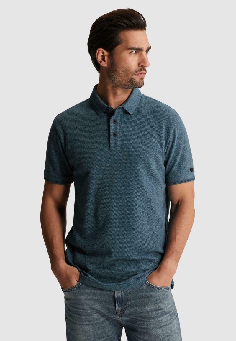 Homme en polo bleu sarcelle à manches courtes et jean, les mains dans les poches, regardant vers la droite sur un fond clair uni.